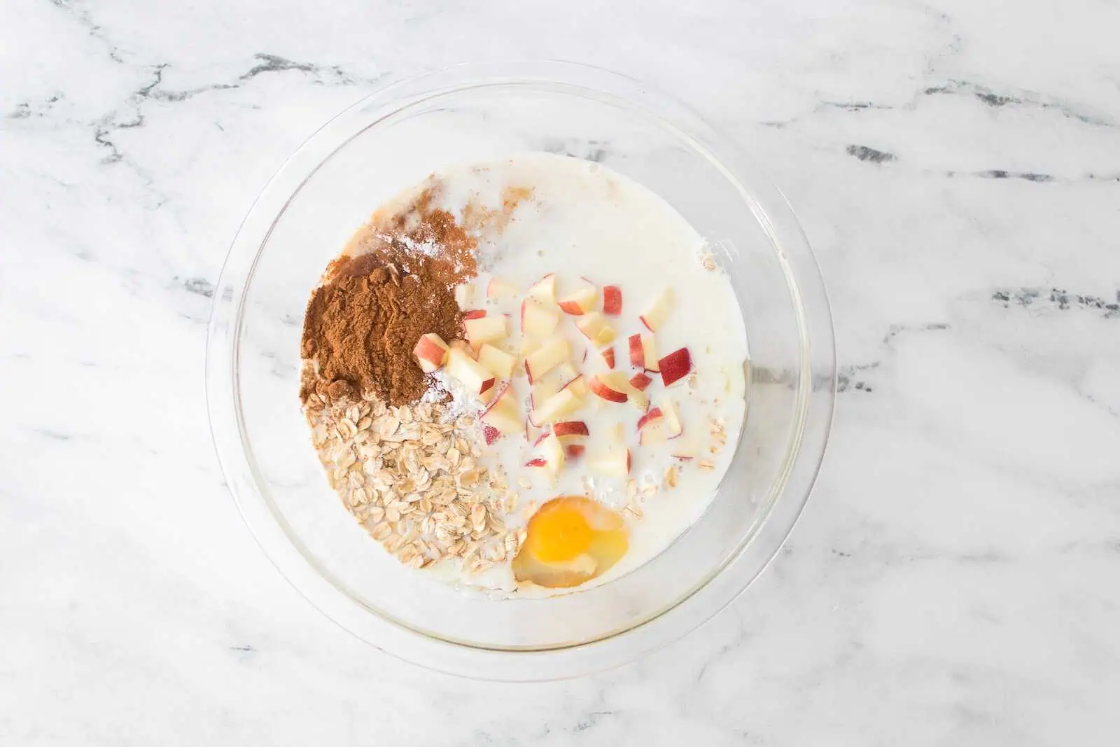 Baking ingredients in a glass bowl, including oats, cinnamon, chopped apples, eggs, and milk, on a marble countertop.
