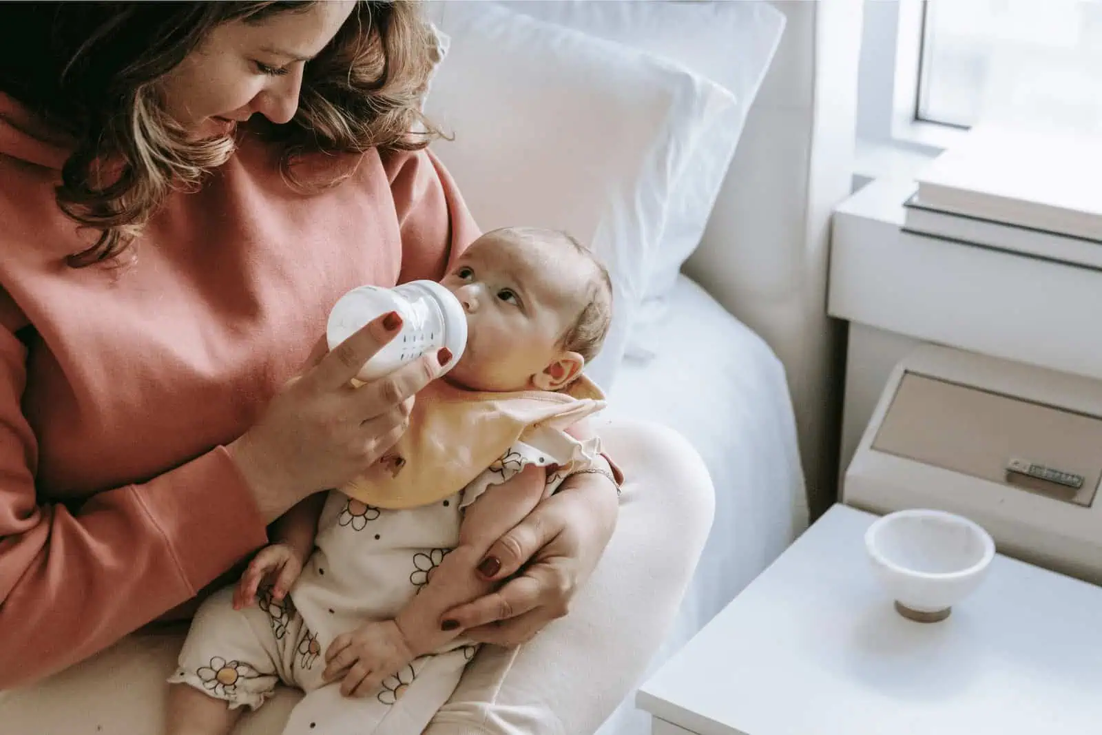 Baby being fed with a bottle by a woman, sitting on a bed in a cozy room.