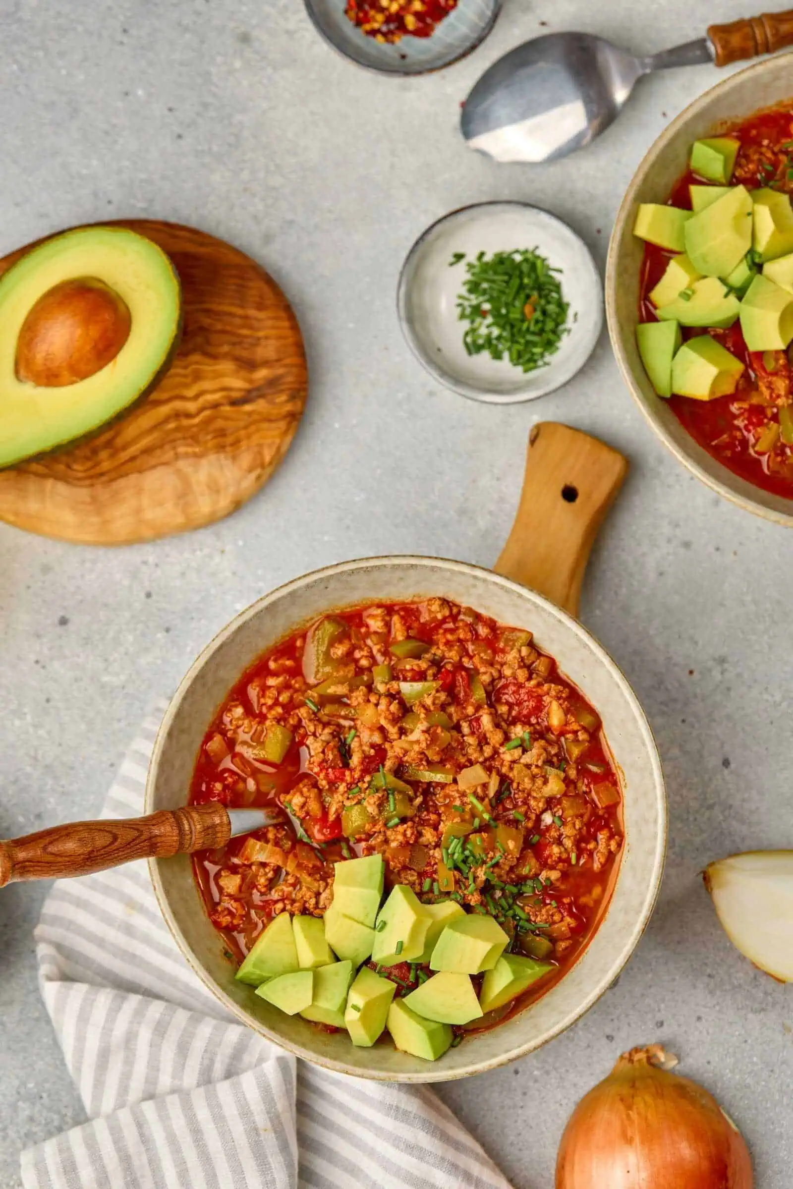 Avocado-topped spicy chili in a beige bowl, fresh ingredients around, healthy comfort food.