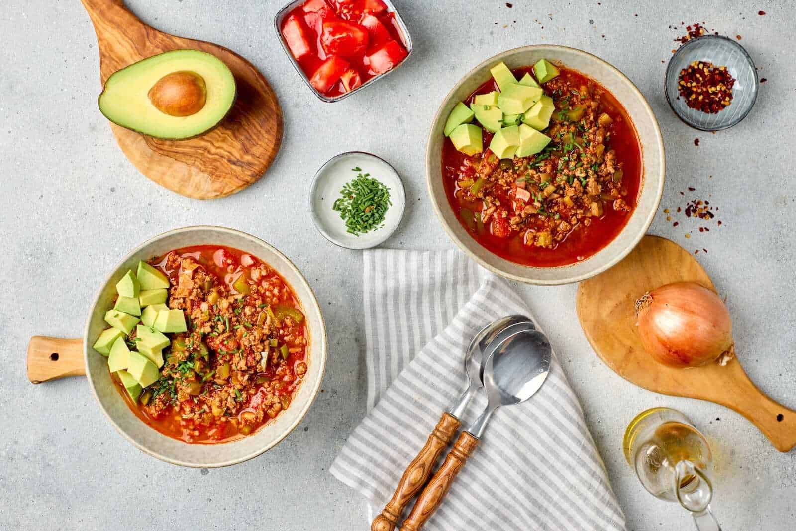 Savory chili with avocado and fresh ingredients on a table featuring bowls, chopped vegetables, and spices.