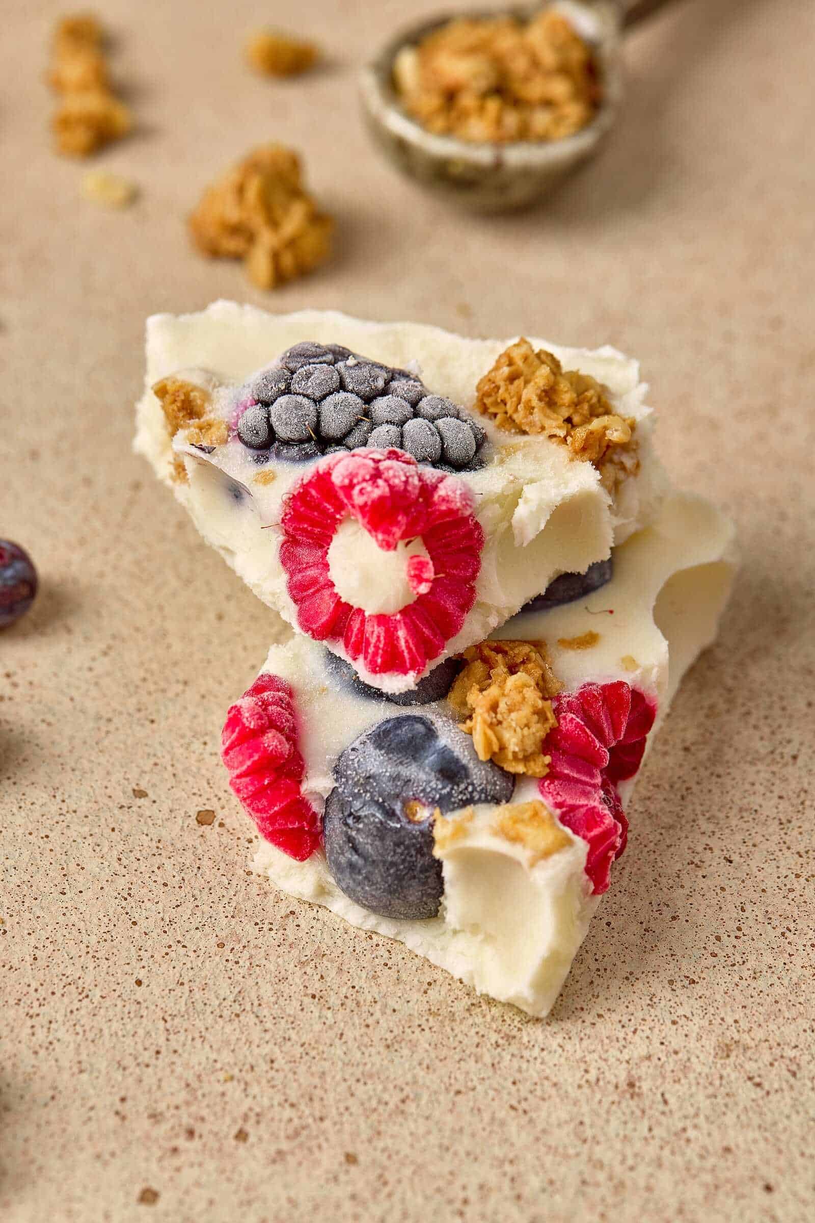 Close-up of berry dessert bar topped with frozen blueberries and raspberries, with granola on a beige surface.