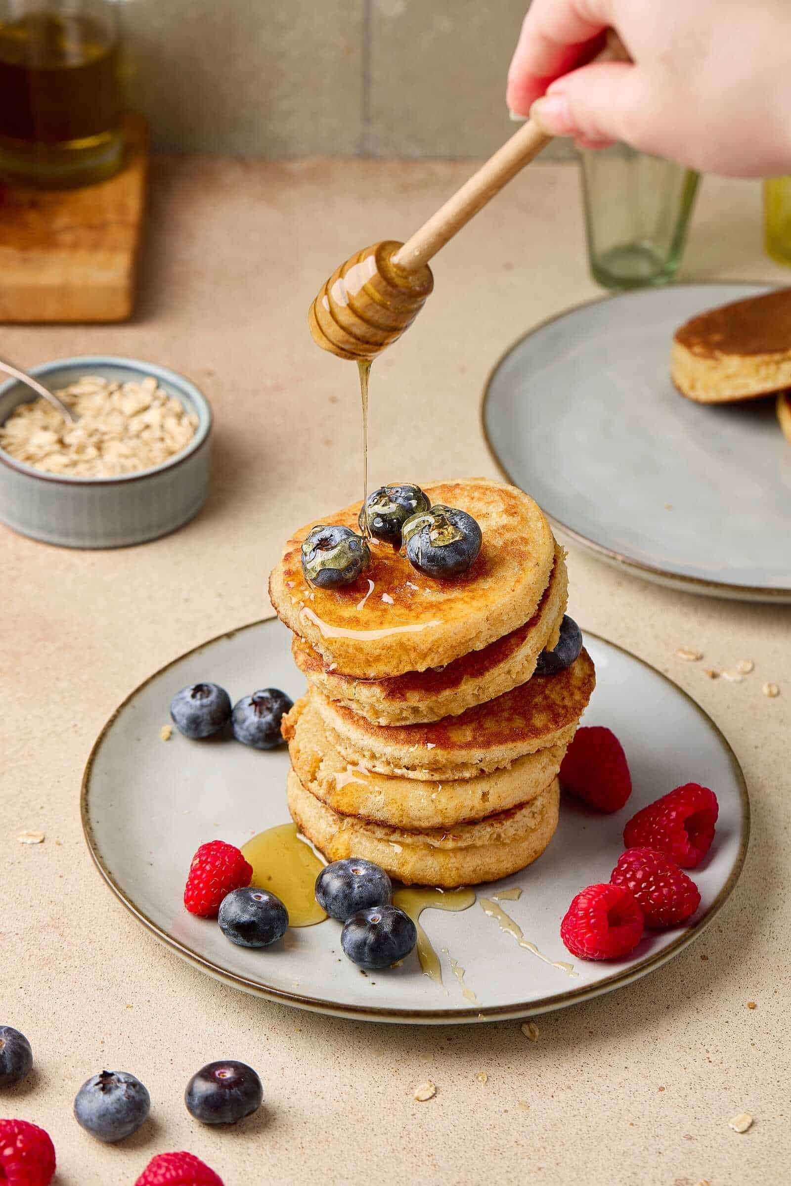 Delicious stack of fluffy pancakes topped with blueberries, raspberries, honey, and oats, perfect for breakfast or brunch.