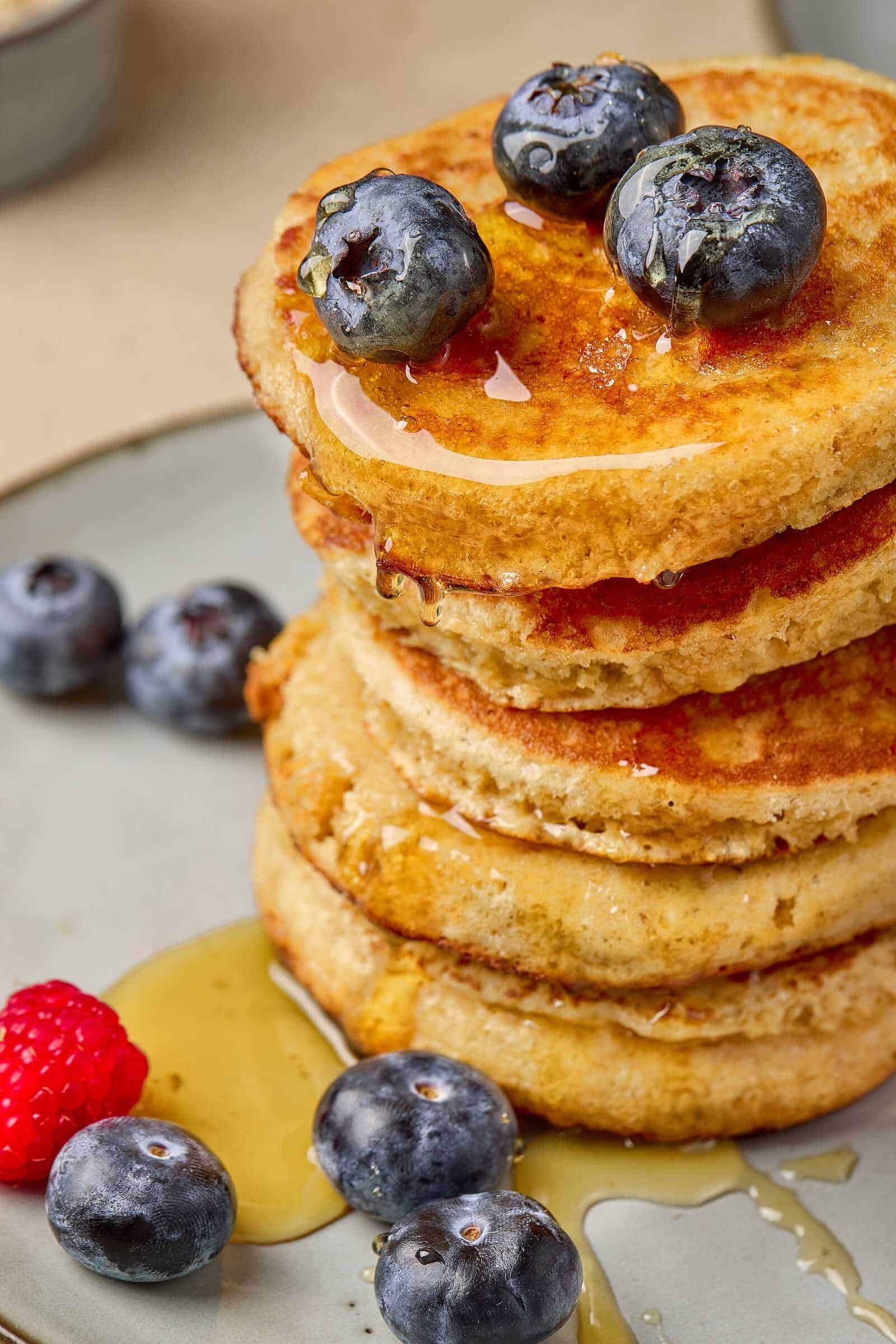 A stack of golden, fluffy blueberry pancakes drizzled with syrup, garnished with fresh blueberries and raspberries, served on a plate.